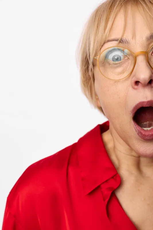 Surprised senior blonde woman in a red shirt and glasses, mouth open and eyes wide on a white studio background. Concept for shock, disbelief, alarm, panic, and stress.