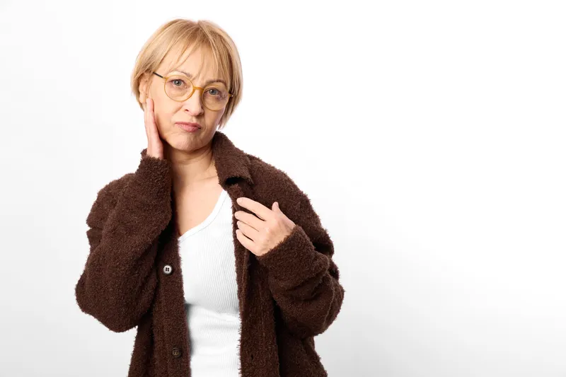 Woman senior with glasses and brown coat, studio portrait, holding collar with hand, concerned and frowning expression, white background, copyspace, casual outerwear