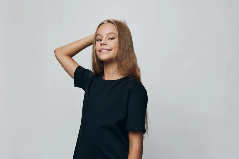 Girl with long hair and black t shirt posing with hand behind head smiling and confident on gray background in studio. Youth, teenager and casual portrait with natural light.