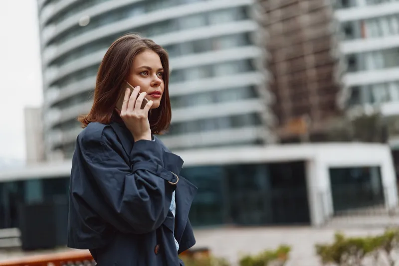 One woman on phone stands on an urban street in a modern city, wearing a dark blue trench coat with brown hair and a focused, calm expression, framed by tall glass buildings.