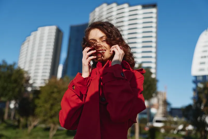 Young woman in a red coat stands outdoors in a city park, wind lifting her hair as she adjusts the coat and smiles softly amid modern buildings.