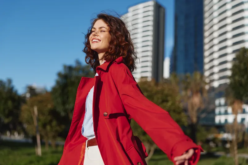 Young woman with curly hair enjoys fresh air outdoors wearing a red coat and white pants in a sunny urban park with modern buildings in the background.