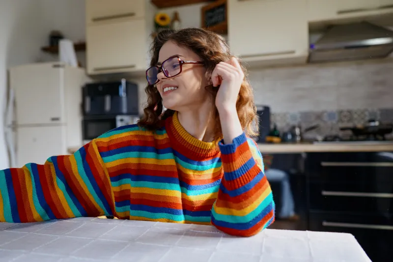 A young woman with curly hair sits at a kitchen table wearing a colorful striped sweater, smiling softly as she rests her arm on the table in a warm, relaxed home moment