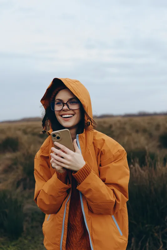 Woman lifestyle outdoors, smiling in an orange hooded jacket, wearing glasses, holding a smartphone, natural field landscape, cloudy sky, warm casual mood