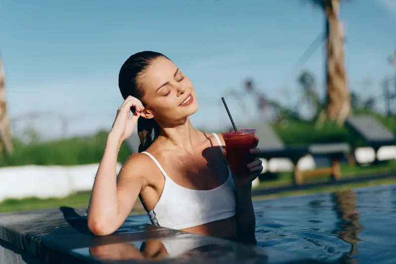 Young woman in a white bikini top relaxes in a pool while enjoying a cold drink on a sunny day with eyes closed and a peaceful smile.