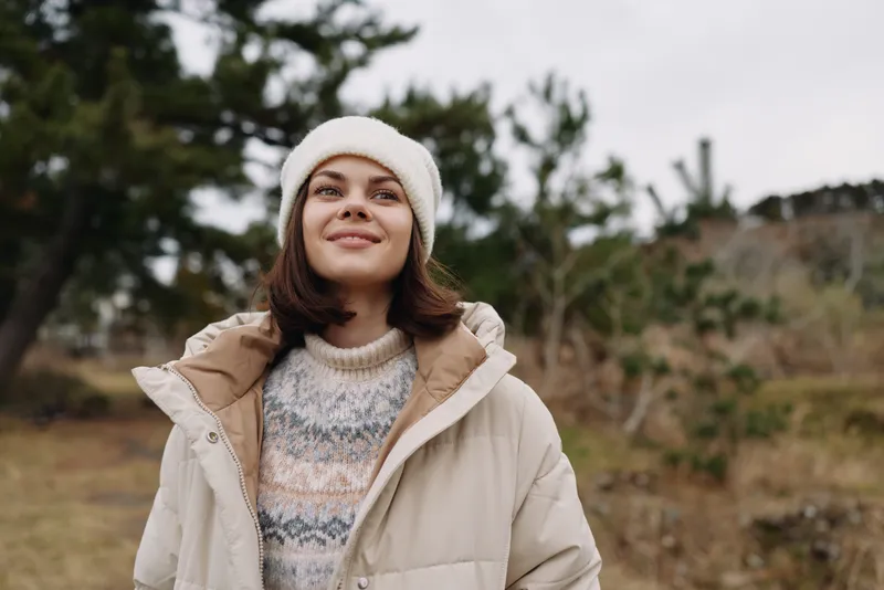 Woman outdoors in a beige coat and knit sweater, smiling in a soft winter mood against a natural landscape of trees and hills, casual fashion and lifestyle portrait for outdoors