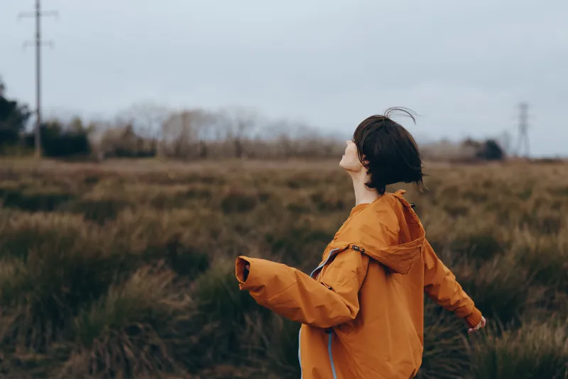 Woman in orange rain jacket with hood, lifestyle outdoors scene in a field, profile view as wind moves her hair, walking in nature meadow, overcast sky soft daylight