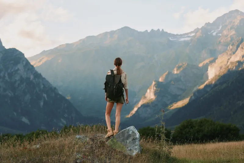 Woman hiker with backpack standing on rock looking at mountain range during sunset, outdoor adventure, nature exploration, casual outfit, peaceful moment