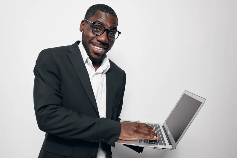African American man with dark skin wearing glasses and black suit smiling while typing on laptop computer isolated on white background in studio.