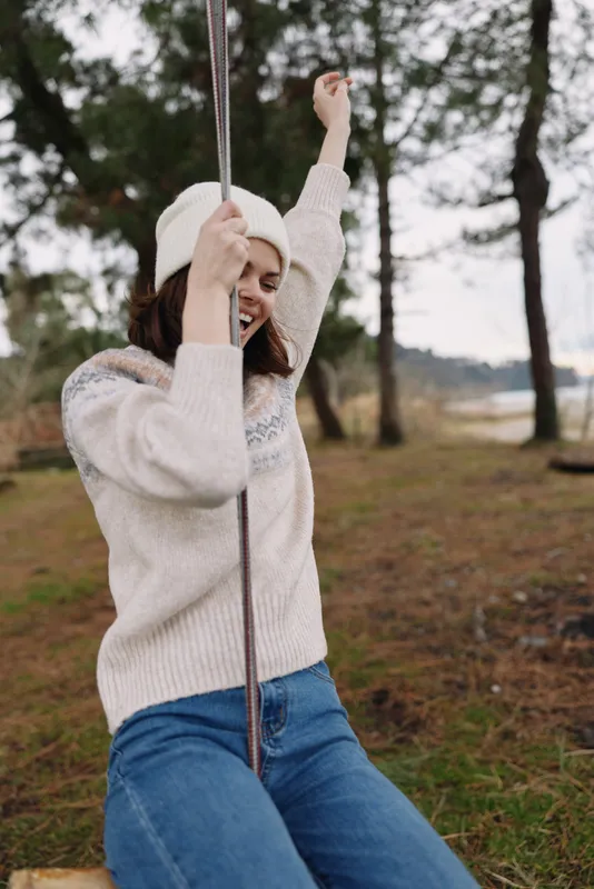 Outdoor swing scene with a woman in a cozy sweater and hat enjoying a carefree moment in a park among trees and grass, conveying relaxed lifestyle and natural charm.