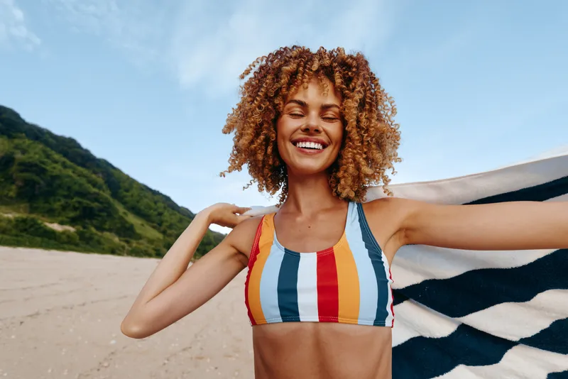 Woman smiling at beach, curly hair, holding towel, outdoor vacation mood, sunlight and mountains background, enjoying summer rest, carefree posture, close view on sandy shore, bright sky