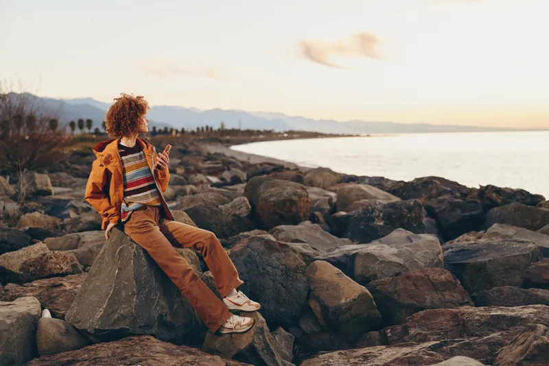Woman lifestyle outdoors on rocky coast at sunset, sitting on stones, wearing orange jacket and backpack, looking at sea horizon, warm sunlight, travel mood, soft waves background