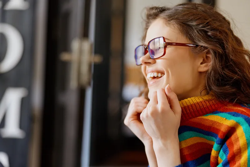 Cheerful woman indoors, wearing a colorful striped sweater and glasses, smiling in a candid moment, warm lighting, home setting, relaxed lifestyle, vibrant tones, cozy atmosphere everyday