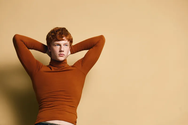 Young man with red hair and brown turtleneck posing with hands behind head against beige background. Casual style, confident expression and relaxed mood in studio portrait.