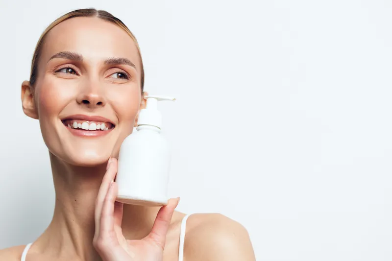 Smiling woman with clear skin holds white cosmetic bottle near face, shows skincare product, demonstrates beauty routine, isolated on white background with natural light and positive expression.