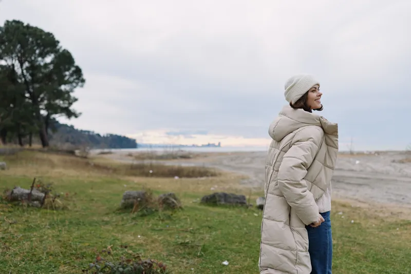 Winter outdoor scene with a person in a long beige coat by a windy coastline, grassy foreground and distant shore, calm mood and open space for travel or nature escape.