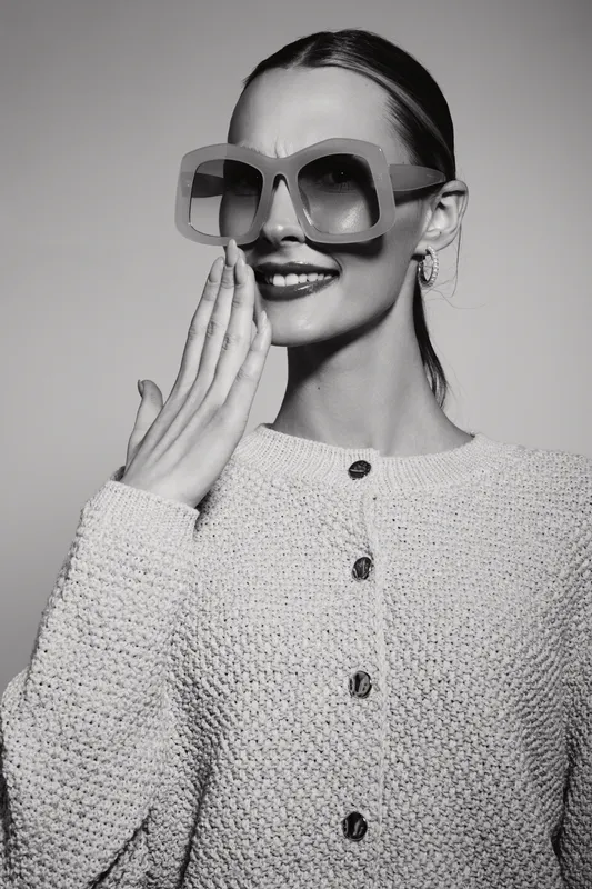 Smiling caucasian woman with light skin wears large sunglasses and knitted sweater, covering mouth with hand. Studio portrait with positive mood.