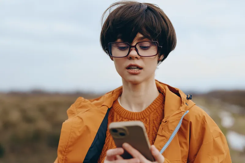 Woman lifestyle portrait outdoors, wearing glasses and a jacket, holding a smartphone and reading messages while traveling in a windy landscape. Soft daylight, casual modern style today.