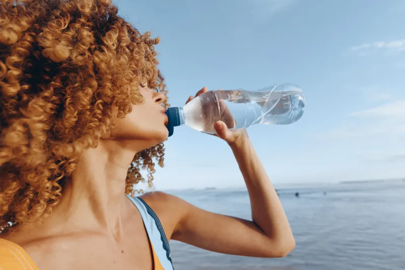 Woman with curly hair drinking water from a clear bottle outdoors by the sea, summer breeze on her face, close side view, bright daylight, hydration refreshment moment, ocean horizon