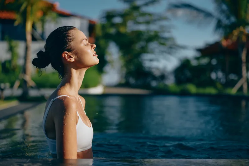 A relaxed woman stands in a sunlit pool, eyes closed, savoring the warm breeze. A casual lifestyle moment that conveys calm, self care, and a peaceful, rejuvenating escape.