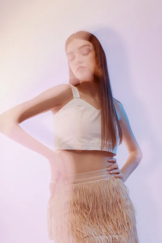 Young woman in a white crop top and fringed skirt poses with eyes closed in a soft pastel light. Concept of beauty, fashion, and elegance with a dreamy mood.