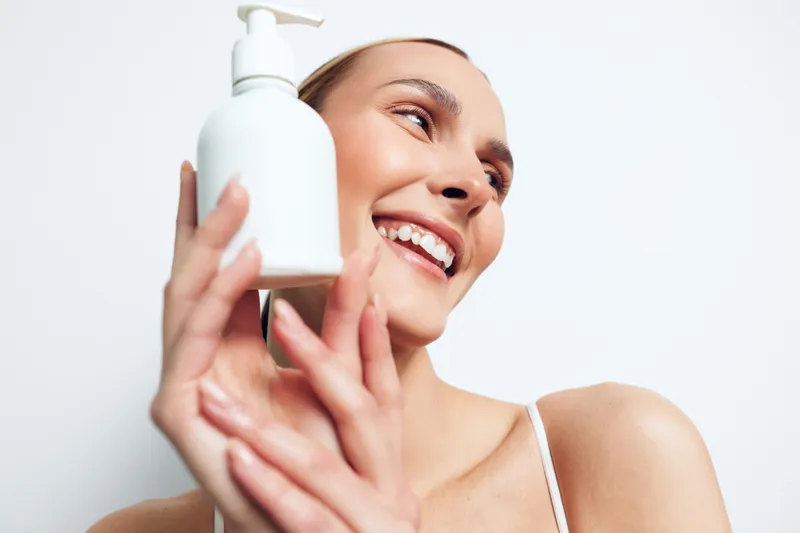 Smiling woman holding white lotion bottle close to face, clear skin, natural makeup, bright background, skincare concept, beauty product, happy expression, soft lighting