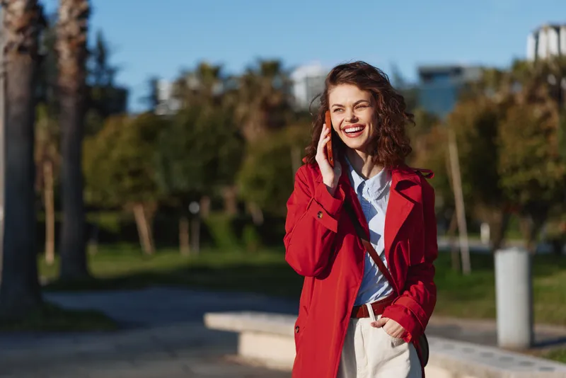Smiling woman with curly hair talks on her phone in a sunny park, wearing a bright red coat over a light shirt and beige pants for a casual outdoor moment.