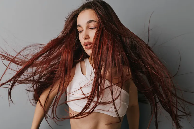 Woman with long red hair wearing white crop top posing with eyes closed and hair flying, studio portrait with neutral background and natural makeup for beauty and fashion.