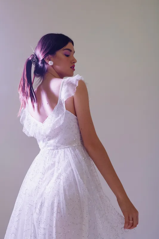 Young woman in a white elegant dress poses with her back turned, showcasing beauty and fashion. Soft lighting highlights delicate textures and feminine style in a studio setting.