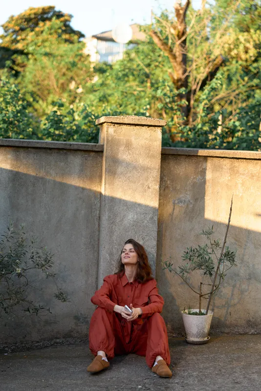 A woman in a red jumpsuit sits on the ground beside a weathered wall in a sunlit courtyard, conveying urban lifestyle, calm solitude, and reflective mood