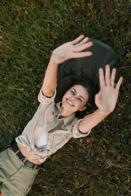 Young woman lying on grass reaches up with both hands, smiling softly in a natural outdoor setting, with a light beige shirt and olive pants under warm sunlight