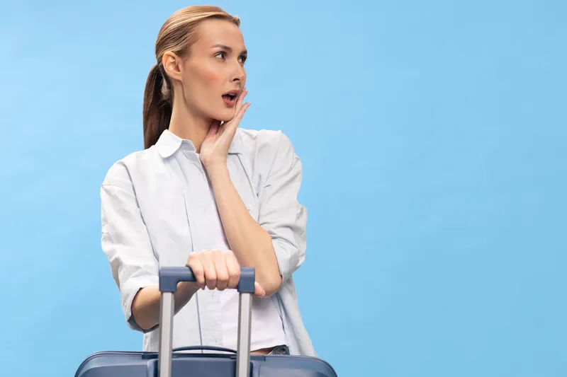 Surprised woman in white shirt holding suitcase handle looks aside with open mouth on blue background. Concept of travel, vacation, shock, and unexpected news for traveler.