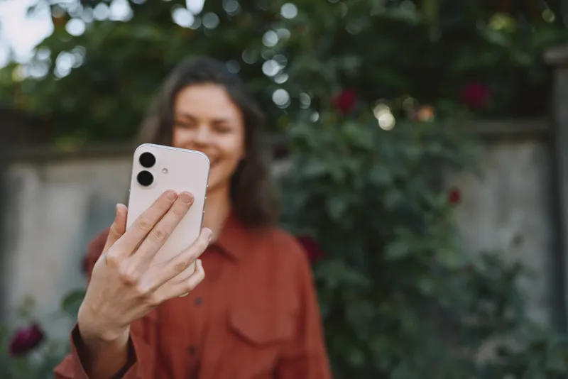 Cheerful woman outdoors taking a selfie with smartphone, casual outfit, sunlit garden background, candid moment, natural colors, vibrant greenery, relaxed mood and self expression