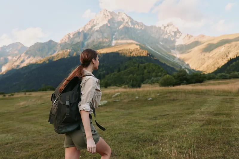 Young woman with backpack hiking in mountains enjoys nature and fresh air during outdoor adventure in sunny summer day with clear sky and green landscape
