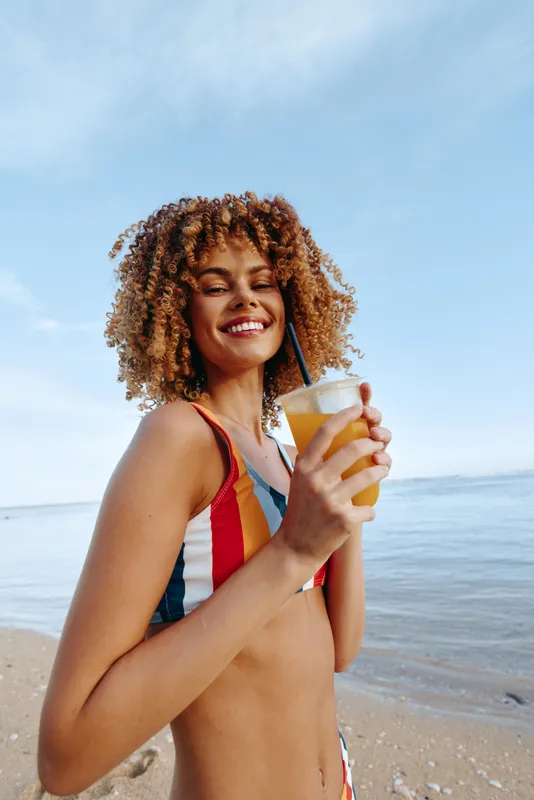 Smiling young woman on a sunny beach holding a cold drink, wearing a colorful swimsuit, enjoying the breeze by the water, vibrant summer mood under bright sky, carefree radiance