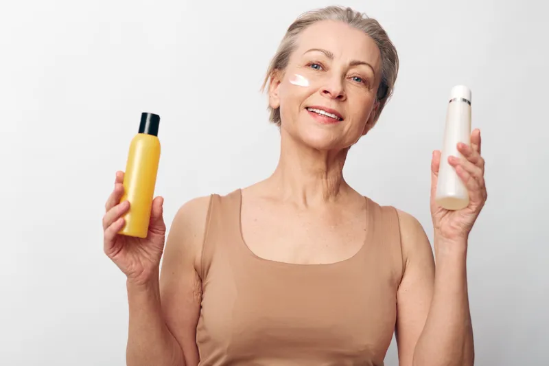Smiling mature woman with short grey hair holds two cosmetic bottles, applies cream on cheek, posing on white background. Concept of skincare, beauty routine, self-care, and aging gracefully.