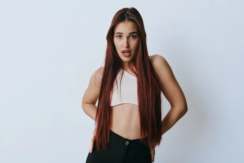 Young woman with long straight hair wearing white crop top and black pants posing with surprised expression on light background in studio. Casual fashion and emotion concept.