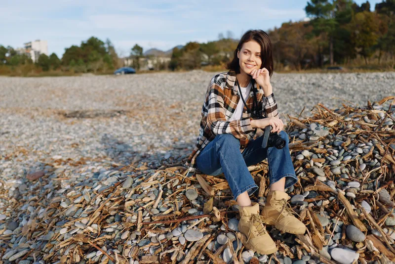 Woman sitting on a pebble beach in casual plaid shirt, jeans and boots, talking on a phone, relaxed mood, autumn light, calm shoreline with distant trees.