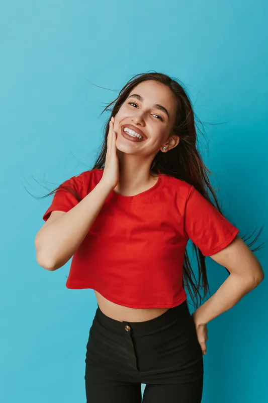 Young woman smiling with braces wearing red crop top and black pants standing against blue background. Happy female model with long hair posing with hand on face and waist.