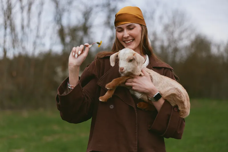 A joyful woman outdoors with a goat kid, cradling the small animal and offering a flower. A warm lifestyle moment highlighting animal companionship and loving care in a rural setting.