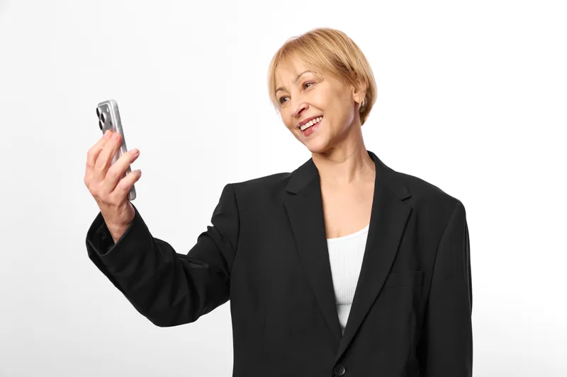 Mature businesswoman smiles holding smartphone up to camera, presenting screen, confident gesture, isolated on clean white studio background, technology communication portrait