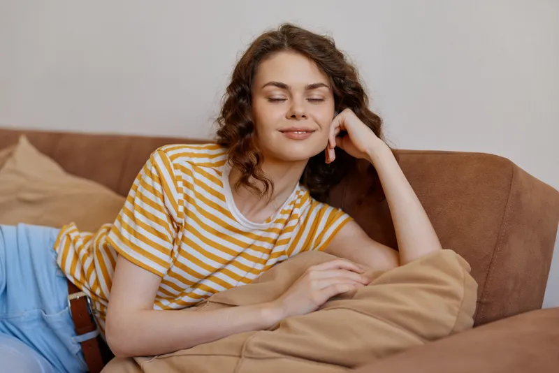 Relaxed young woman with curly hair rests on a couch at home, wearing a yellow striped tshirt, eyes closed with a peaceful smile in a cozy living room, surrounded by cushions and warm natural light