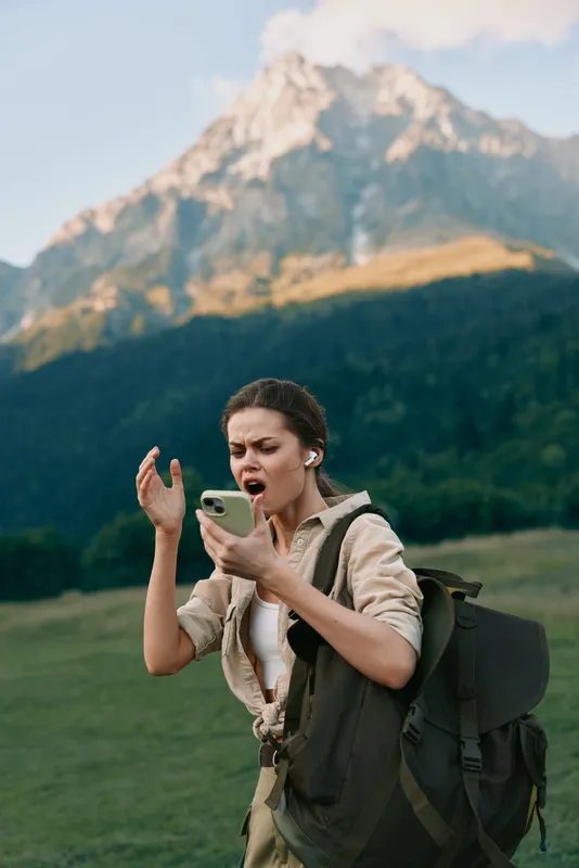 Hiking woman with a backpack checks her phone in a mountain meadow, surprised expression. Outdoor lifestyle, travel vibe, and nature exploration. Fresh air and wide skies.