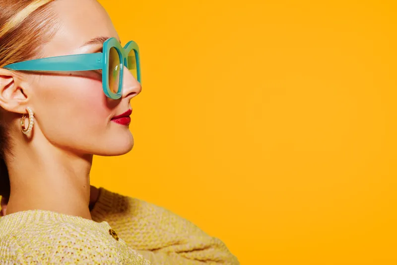 Profile of a stylish woman wearing turquoise sunglasses and gold earrings against a bright yellow background. Concept of fashion, summer, and vibrant lifestyle.