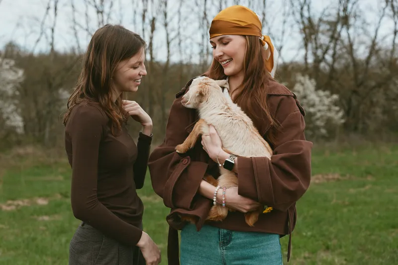 Two young women enjoy a spring day outdoors as one holds a small goat while they smile and share a joyful moment in casual clothing in a natural countryside setting.