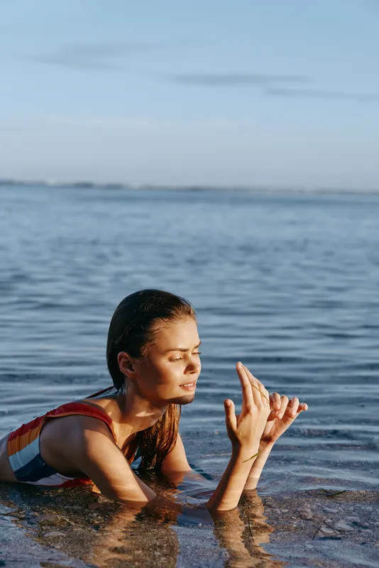 Smiling girl in shallow ocean water, playful hand gesture, summer beach outdoors, gentle waves and blue sky, sunlit light, childhood joy, natural colors, calm horizon, relaxed posture