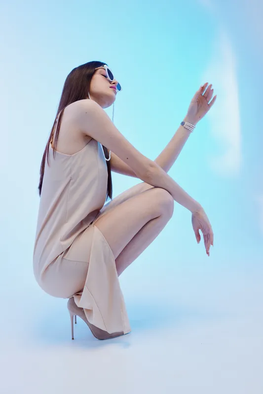 Stylish woman in a beige dress and high heels poses in a squatting position against a blue background. Concept of beauty, fashion, elegance, and modern style.