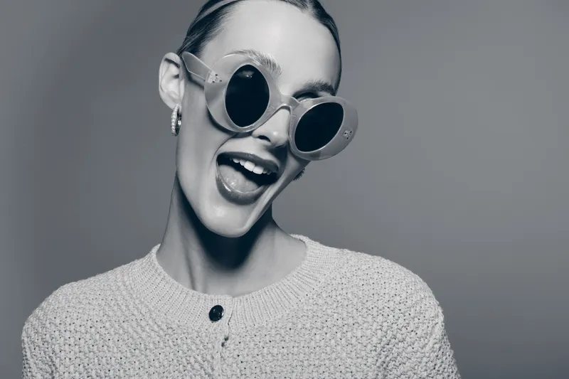 Happy young white woman with fair skin smiles widely wearing round sunglasses and knitted sweater on neutral background. Concept of joy, fashion, and youthful style in studio lighting.