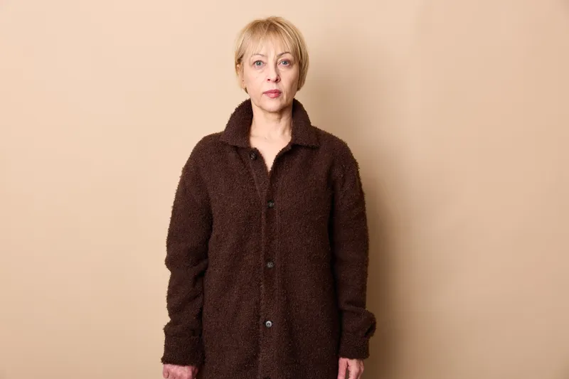 elderly woman coat portrait standing beige studio fashion serious age, senior in dark outerwear on plain background, neutral face expression, soft studio light, calm winter style