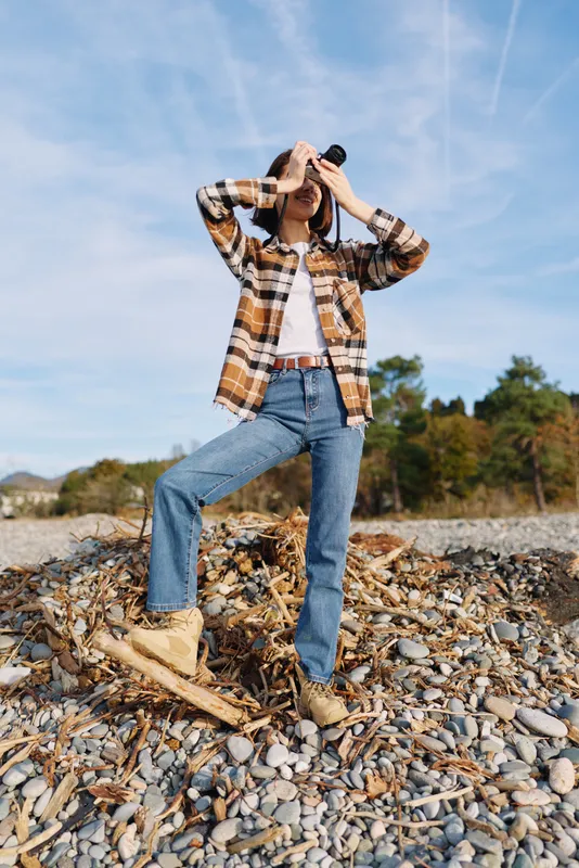 Photographer in casual wear on a rocky beach, using a camera to capture a moment. Plaid shirt, jeans and boots frame the scene as a traveler explores coastal terrain under clear blue sky.
